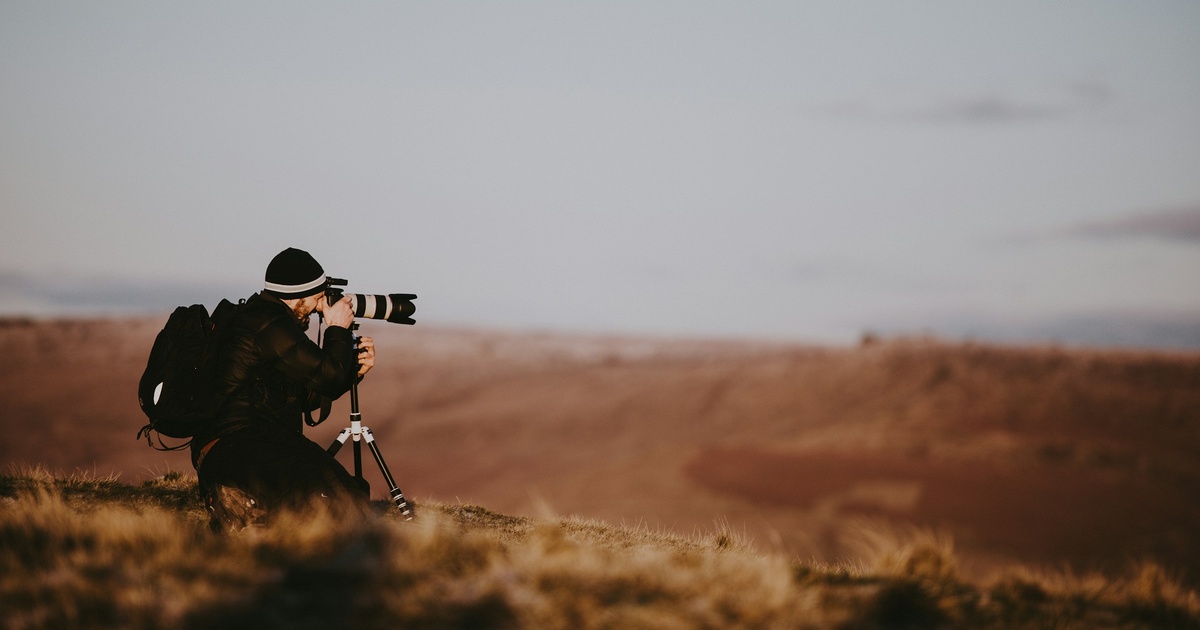 A photographer kneels behind a long lens on a tripod on an open hillside, in low, warm light.